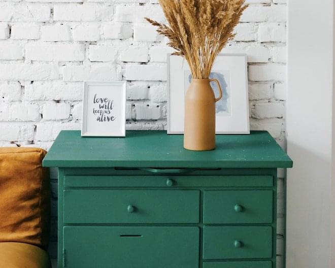 A green dresser against a brick wall painted white with two frames and a vase.